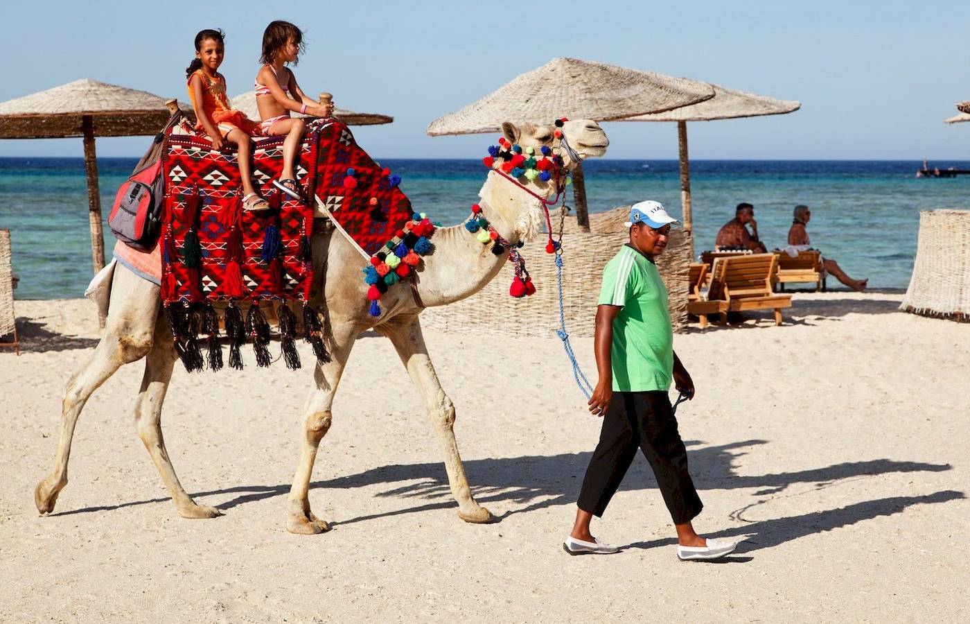 Zwei Kinder reiten auf einem Kamel am Strand, ein Mann führt das Kamel.