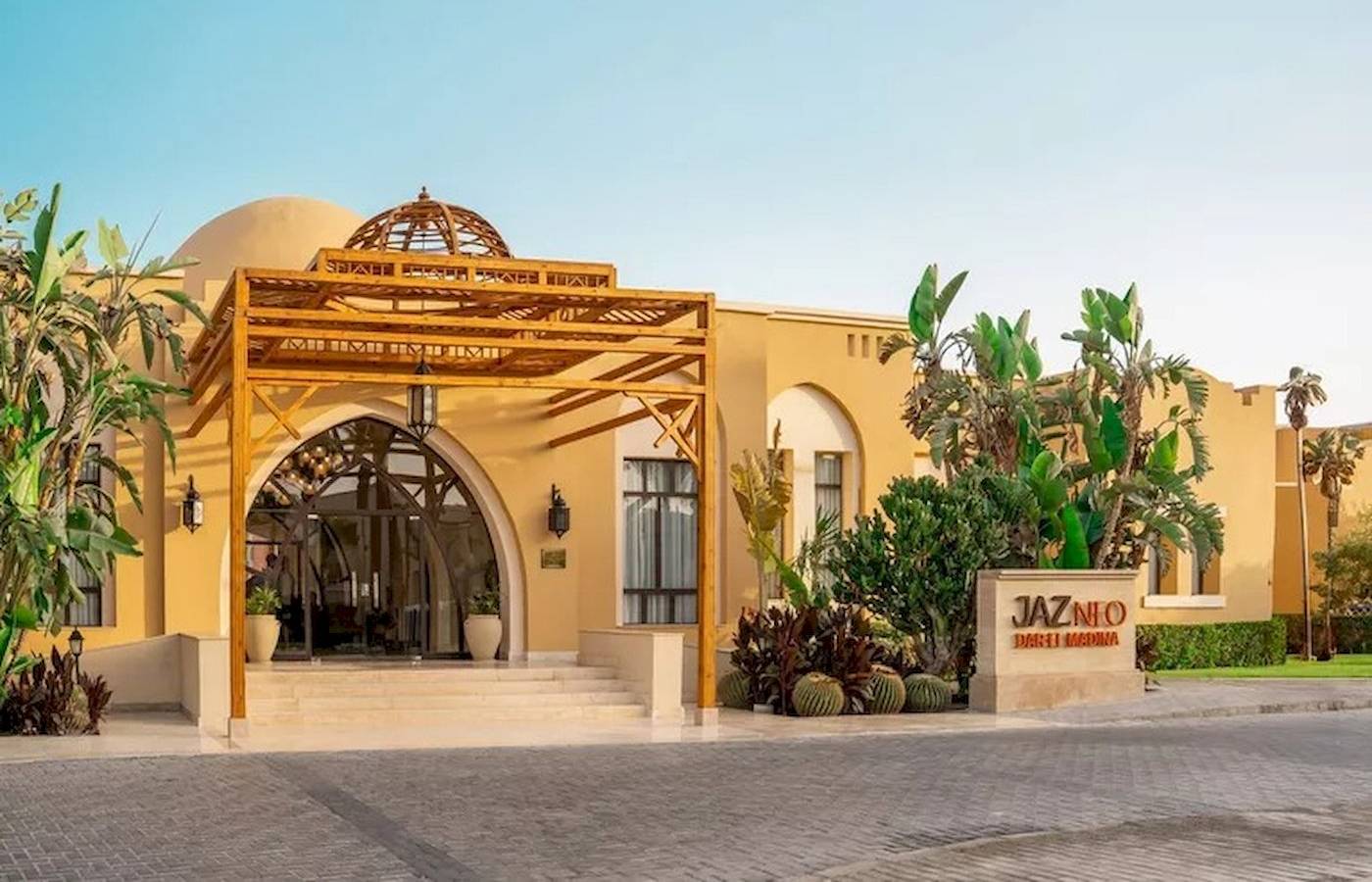 Außenansicht des Jaz Neo Dar El Madina Hotels mit gelben Wänden, architektonischem Eingang und grünen Pflanzen davor.