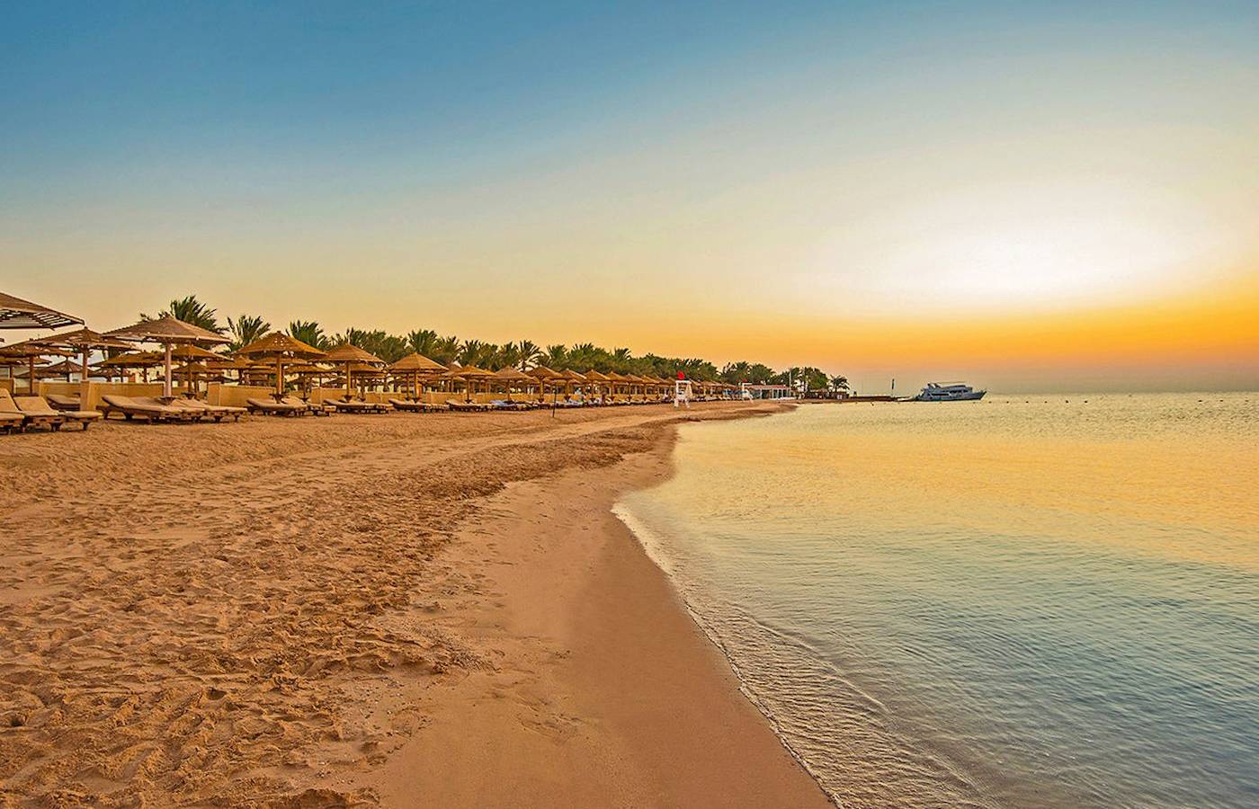 Sonnenuntergang am Strand des SUNRISE Royal Makadi Resort mit leeren Sonnenliegen und einem Boot im Wasser.