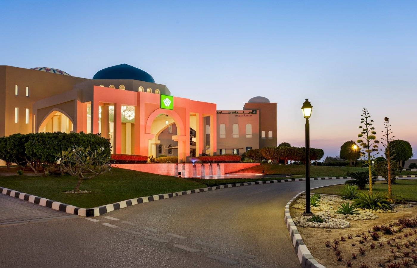 Aussenansicht des Wyndham Garden Salalah Mirbat Hotels bei Sonnenuntergang mit Brunnen und Lampen.