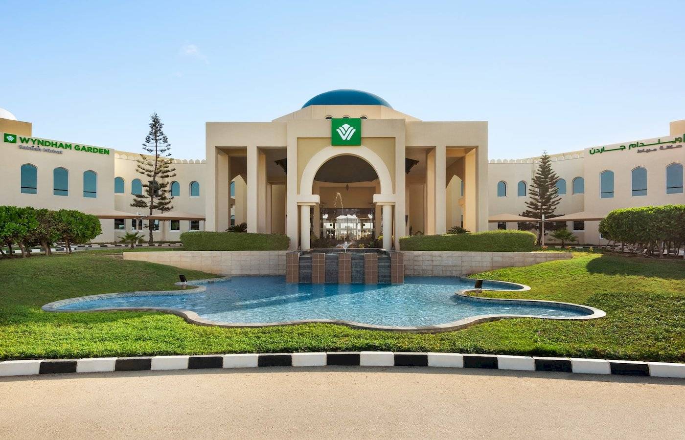 Außenansicht des Wyndham Garden Salalah Mirbat Hotels mit Brunnen und Rasenfläche.
