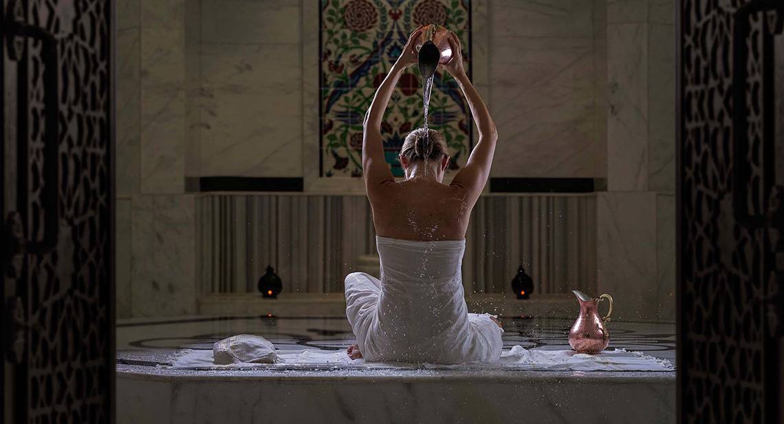 Frau im Bademantel gießt Wasser über ihren Kopf in einem Spa-Bereich