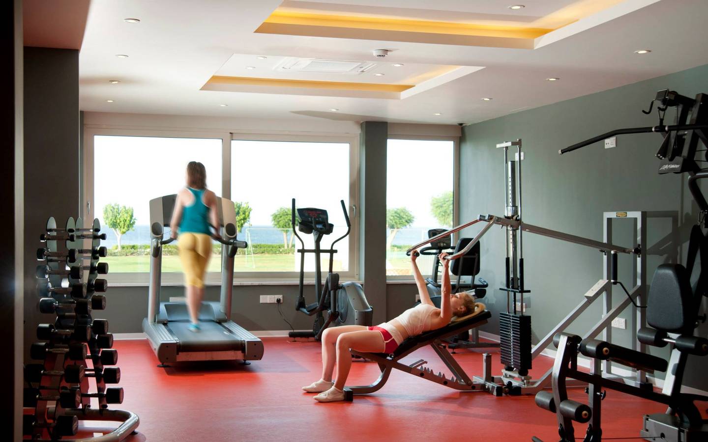 Fitnessraum im Pernera Beach Hotel mit einer Frau auf einer Bankbank und einer anderen Frau auf einem Laufband.