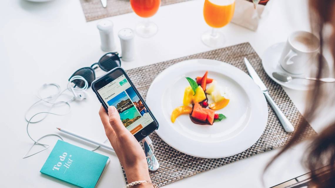 Eine Frau hält ein Smartphone mit der Website von Iberostar Waves Alcudia Park vor einem Frühstückstisch.