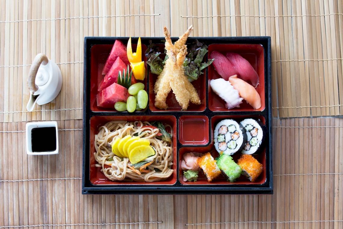 Asiatisches Essen in Schalen auf einer Bambusmatte mit verschiedenen Gerichten wie Sushi, Nudeln und Früchten.