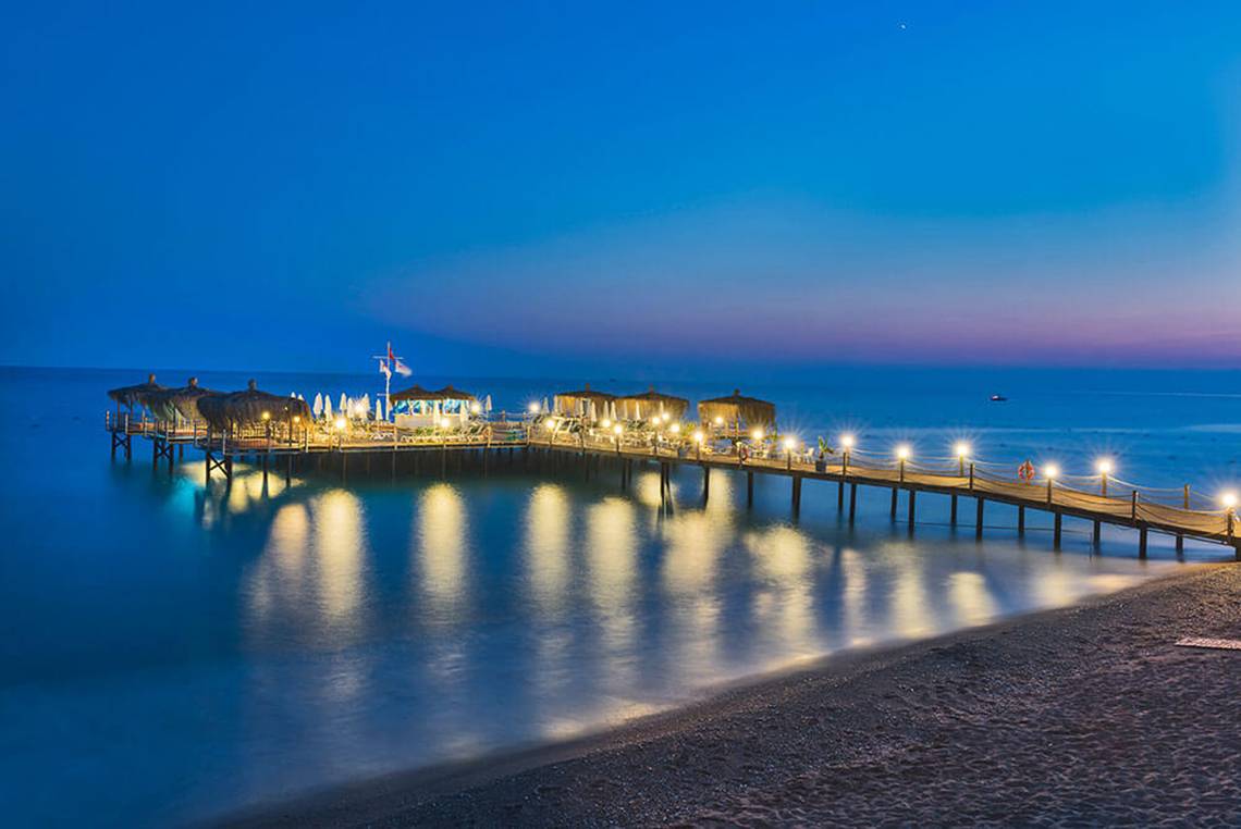 Der Club Nena Pier bei Nacht mit beleuchteten Pavillons und einem ruhigen Meer.