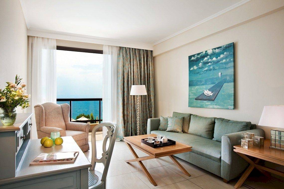 Wohnzimmer im Ikos Oceania Hotel mit Sofa, Tisch, Blumenvase und Meerblick.