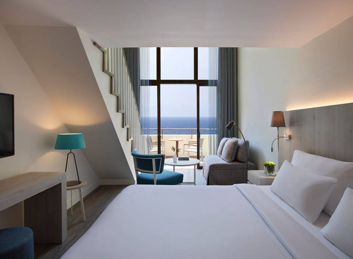 Zimmer im Rhodes Bay Hotel & Spa mit Meerblick, inklusive Bett, Sofa, Tisch und Fenster mit Außenterrasse.