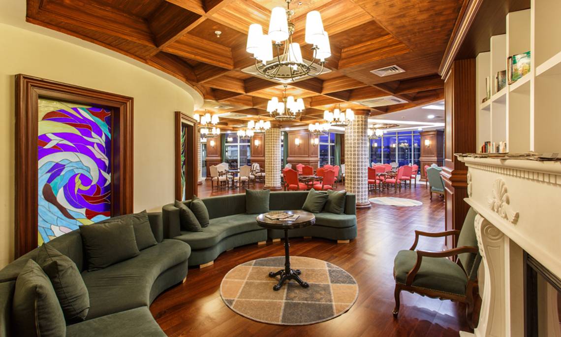 Innenbereich des Sherwood Exclusive Lara Hotels mit grüner Couch, Tisch und farbiger Glasmalerei.
