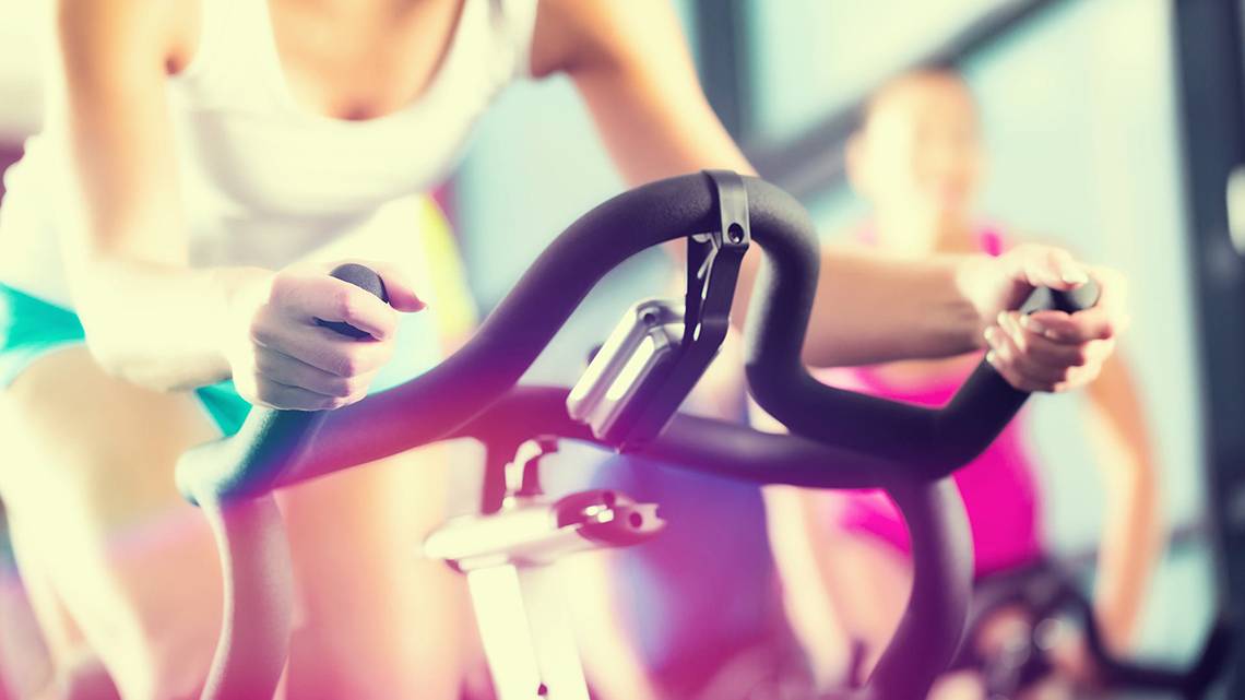 Frau im Fitnessstudio auf einem Fahrrad, während eine weitere Person im Hintergrund ebenfalls fährt.
