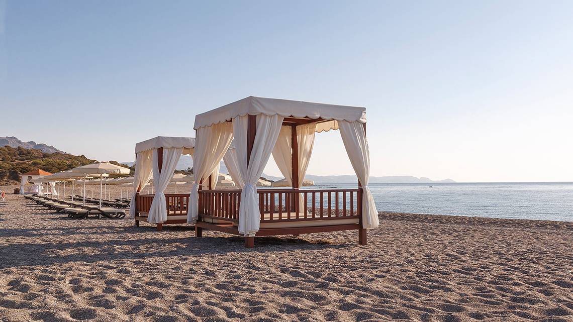 Zwei Strandbetten mit weißen Vorhängen und Sonnenschirmen auf dem Strand des Boutique 5 Hotels & Spa.