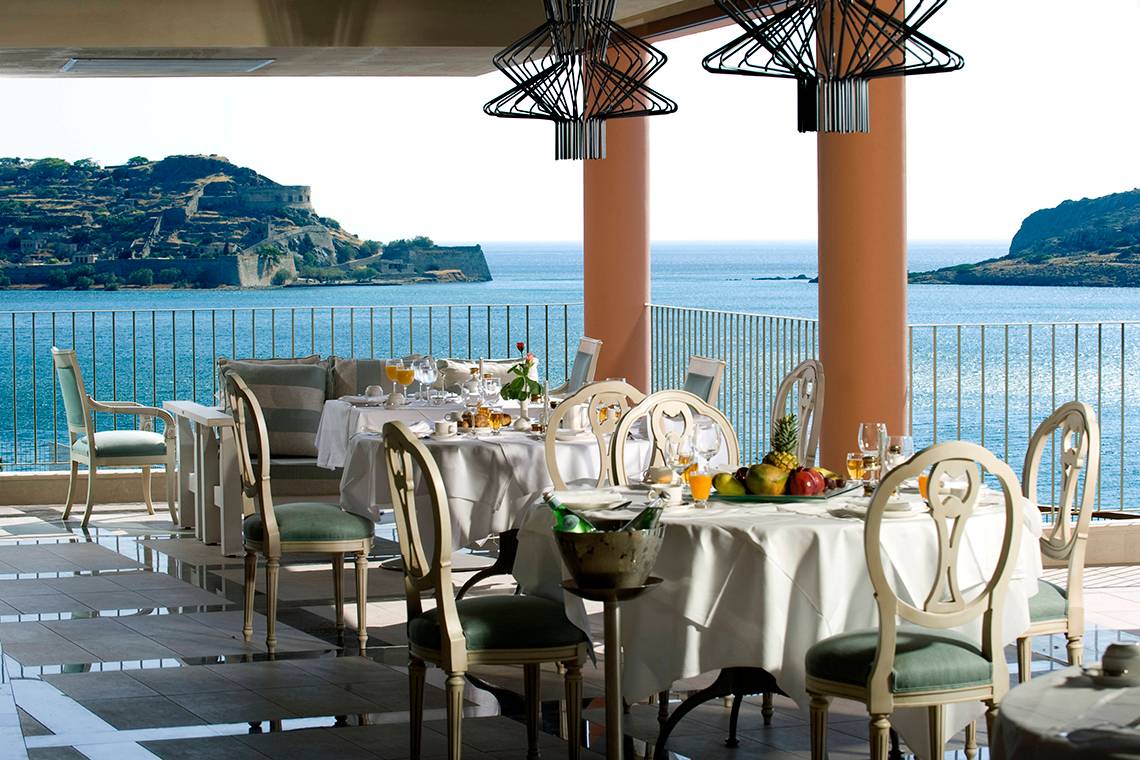 Terrasse mit Meerblick im Domes of Elounda, Autograph Collection mit gedeckten Tischen und Früchten.
