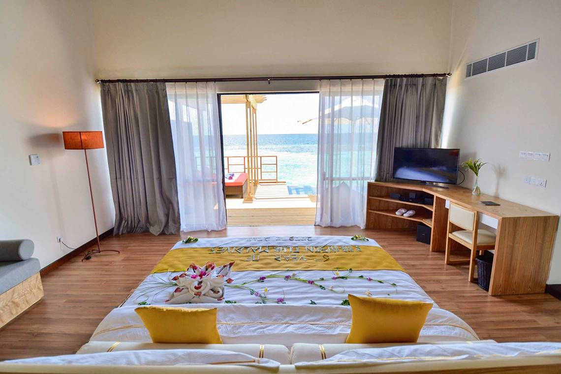 Innenansicht eines Zimmers im Robinson Maldives mit Meerblick, Bett mit Deko, Fernseher und offener Tür zum Balkon.
