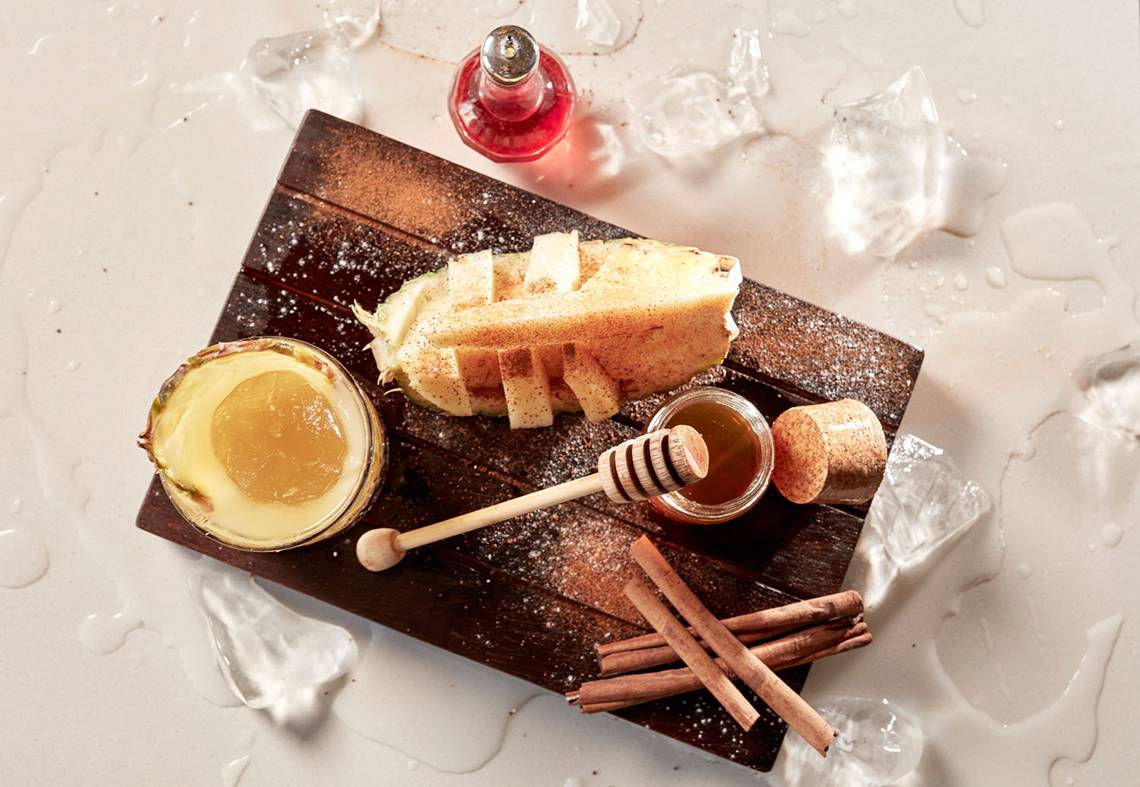 Avra Imperial Essenz Cocktail auf einem Holzbrett mit Eiswürfeln, Zimtstangen und Honig.