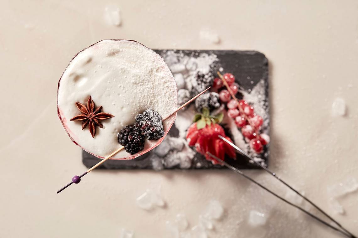 Ein Cocktail mit weißer Creme, Zimt und Beeren auf einem schwarzen Teller.