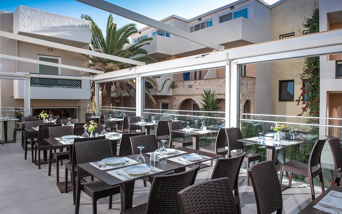 Terrasse des Odyssia Beach Hotels mit Tischen und Stühlen unter Glasdächern.