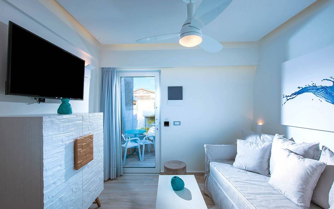 Wohnzimmer des Odyssia Beach Hotels mit Fernseher, Sofa, Tisch und Terrassentür.