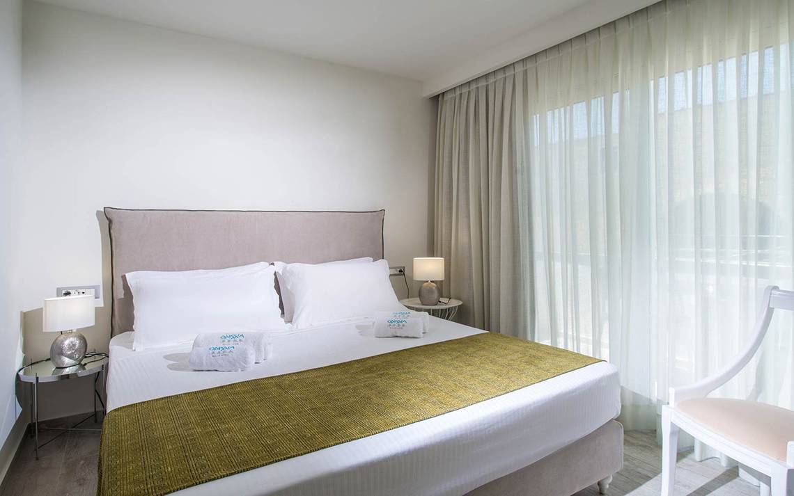 Schlafzimmer im Odyssia Beach Hotel mit Bett, Kissen, Lampen und Fenstern.