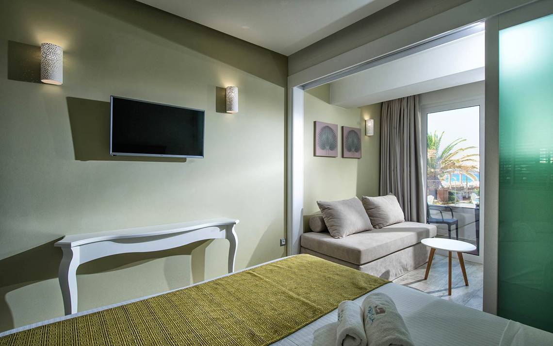 Innenansicht eines Zimmers im Odyssia Beach Hotel mit Fernseher, Couch und Tisch.