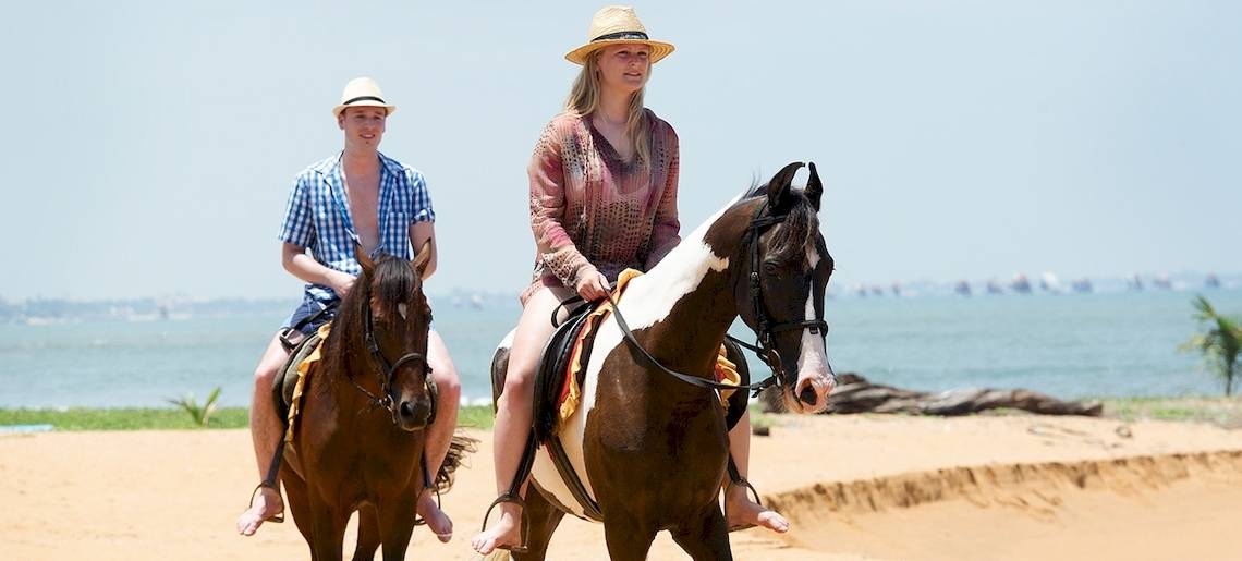 Zwei Leute reiten Pferde am Strand, mit dem Meer im Hintergrund.