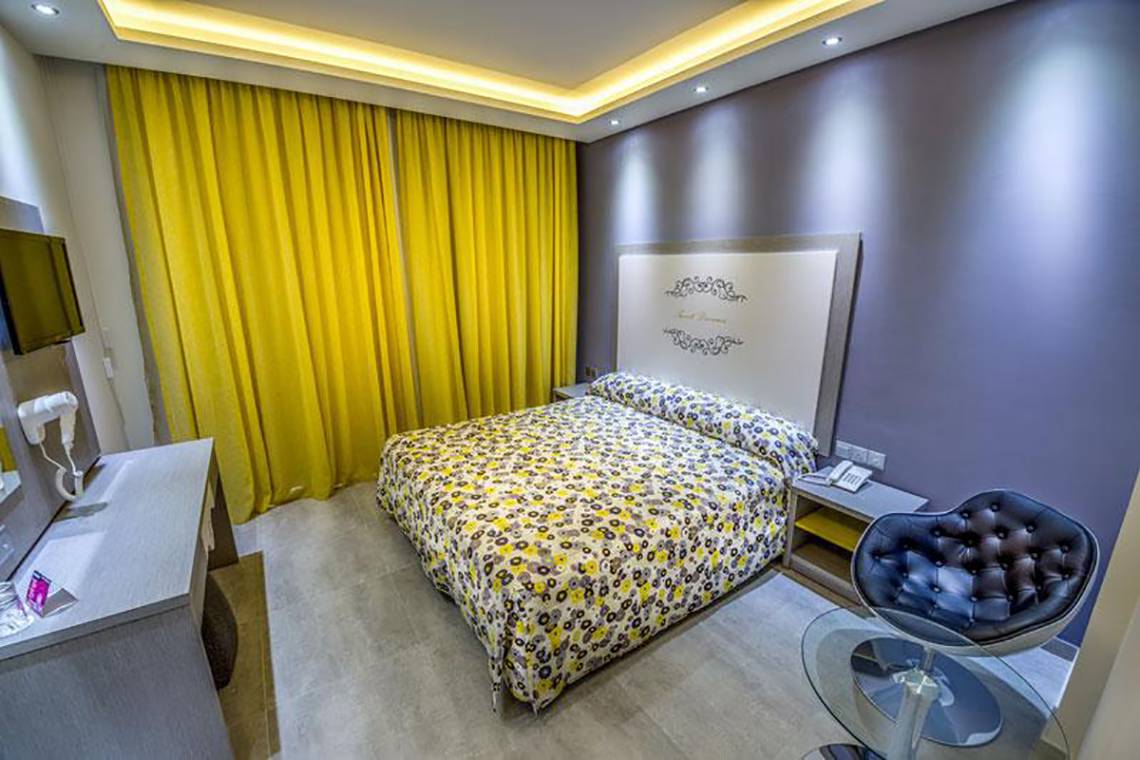 Innenansicht eines Hotelzimmers im Stamatia Hotel mit Bett, Nachttischen und gelben Vorhängen.