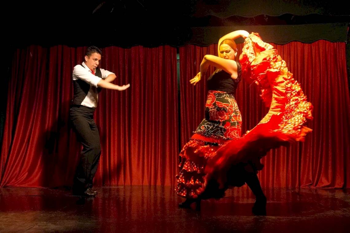Ein Mann und eine Frau tanzen Flamenco auf einer Bühne mit roten Vorhängen im Hintergrund.