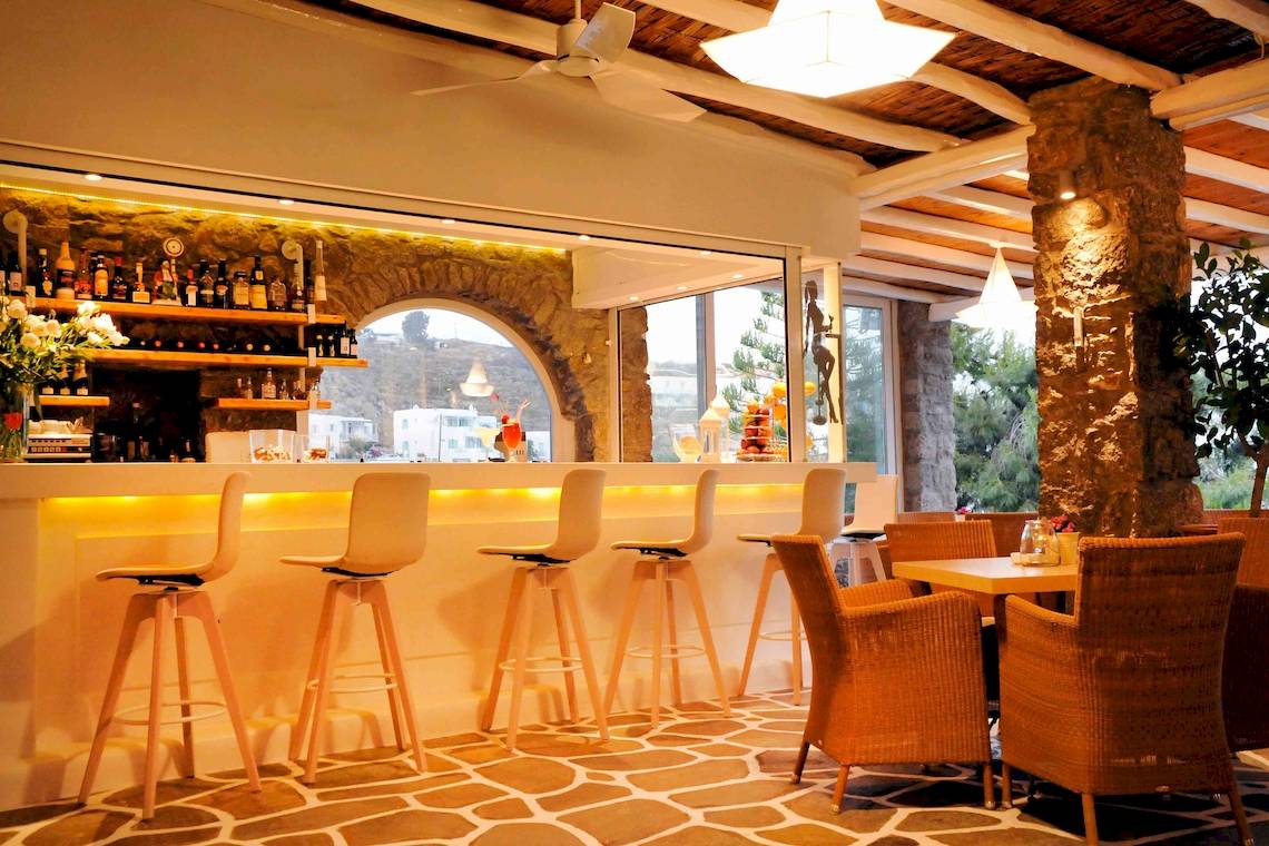 Innenansicht der Bar im Kamari Hotel mit Barhockern, Tischen und Blick auf den Garten.