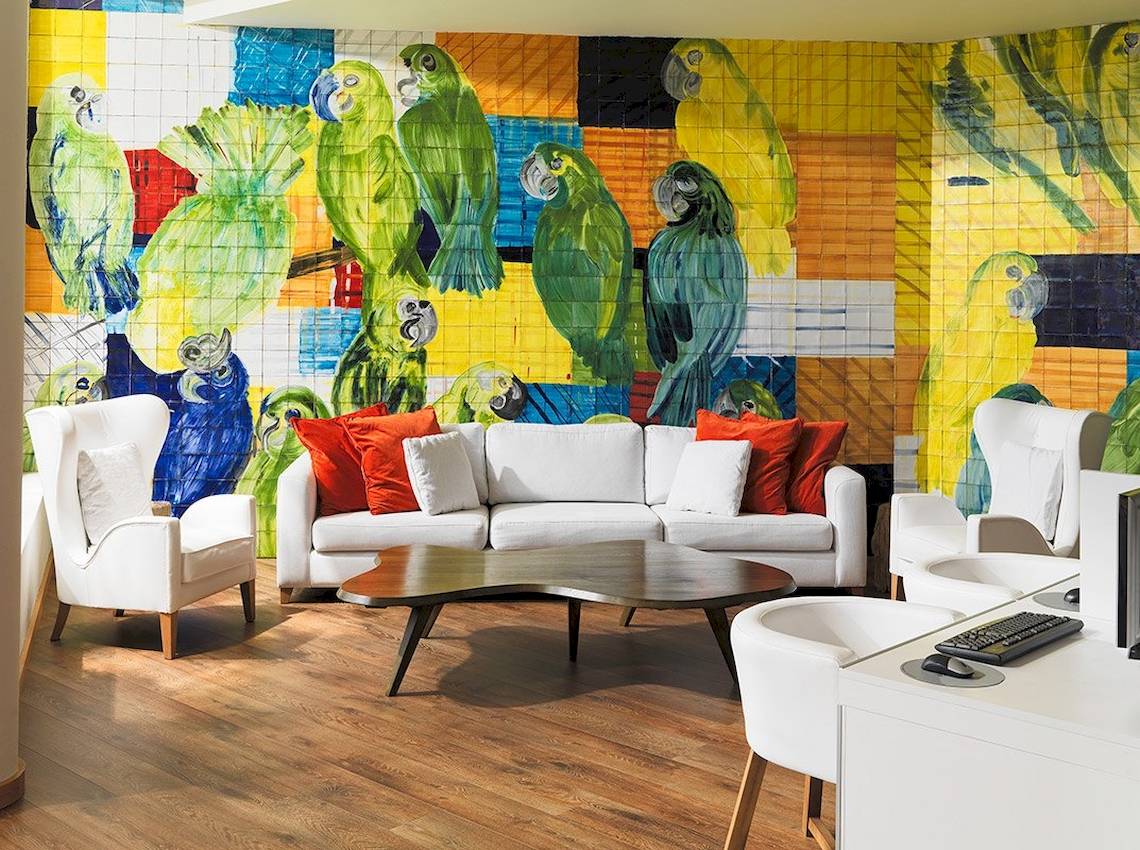 Wohnzimmer mit weißem Sofa, rote Kissen und bunter Wandmalerei von Papageien.