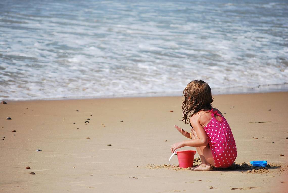 Ein kleines Maedchen in einem roten Badeanzug mit Punkten spielt am Strand mit einem roten Eimer