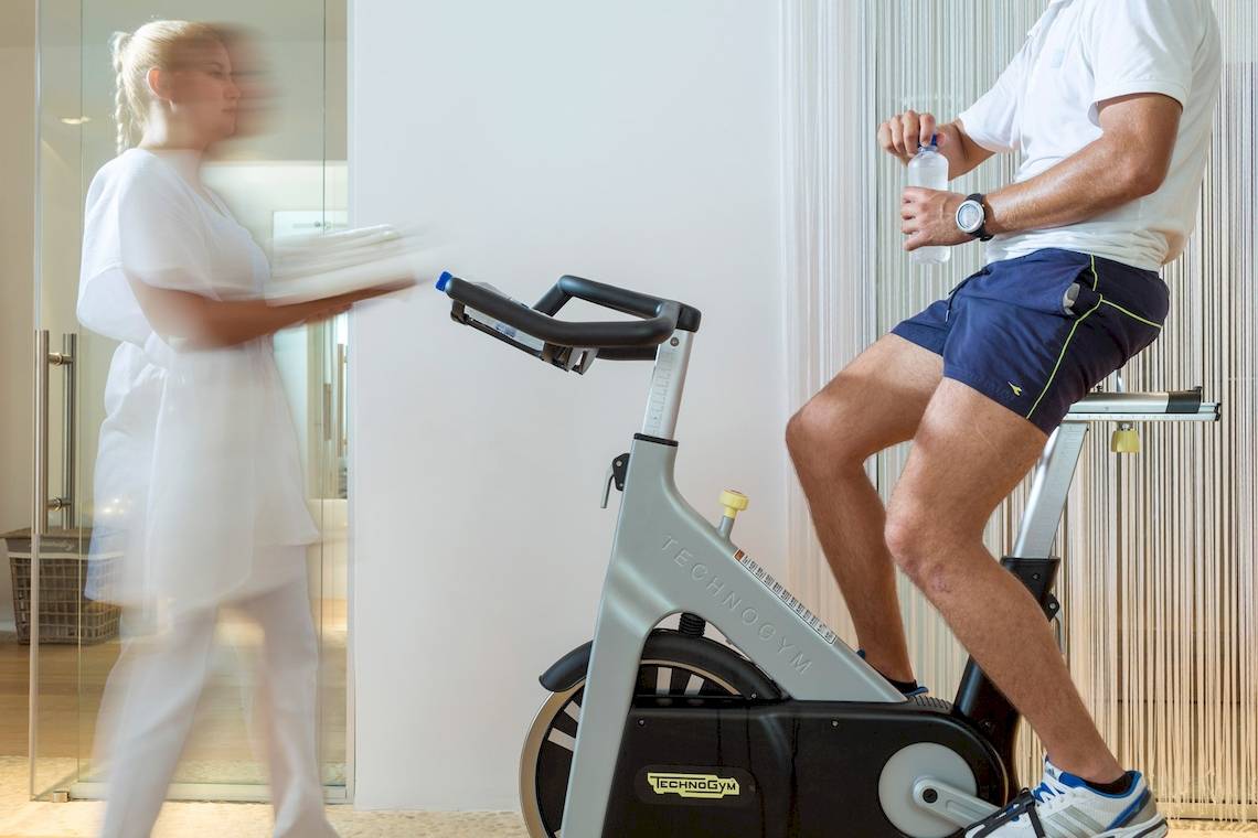 Fitnesstrainer auf einem Technogym-Fahrrad im Katikies Chromata Santorini, eine Trainerin hält Handtücher in der Hand.