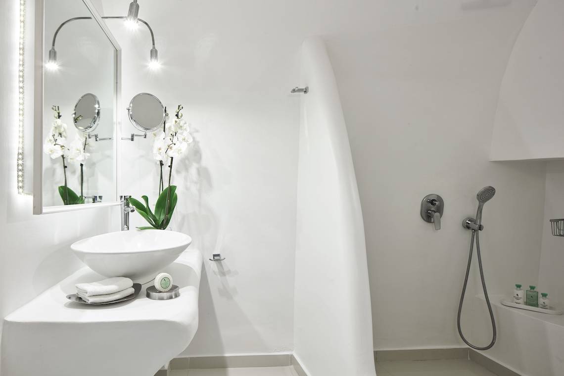 Badezimmer im Katikies Chromata Hotel auf Santorini mit einem weißen Waschbecken, Spiegel und Dusche.