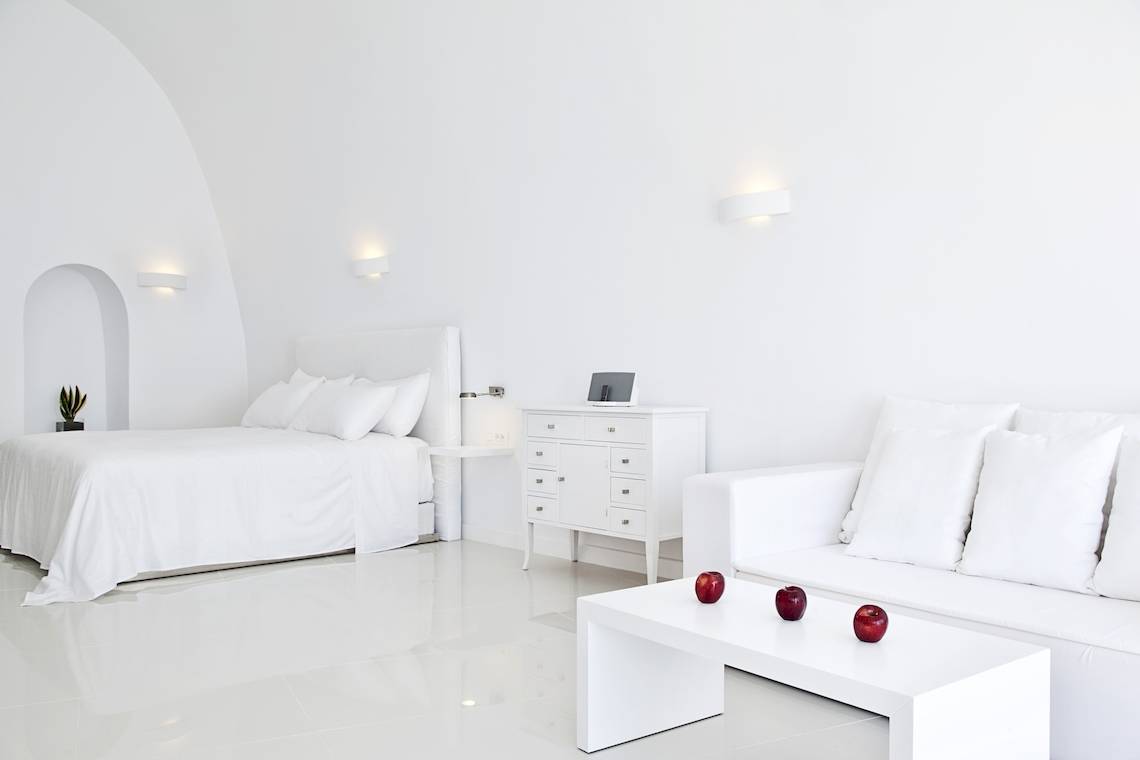 Weißes Schlafzimmer im Katikies Chromata Hotel auf Santorini mit Bett, Nachttisch, Kommode und Früchten auf dem Tisch.