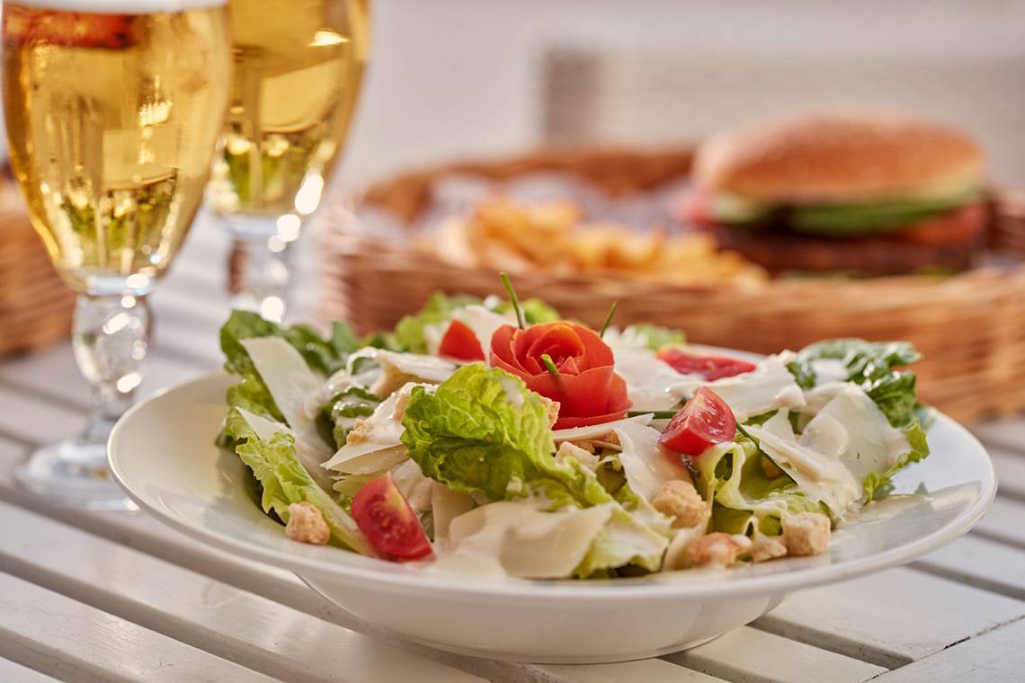 Ein Teller mit einer Salatmischung und einer Rose, mit zwei vollen Weingläsern und einem Korb mit Pommes und Burgern im Hintergrund.