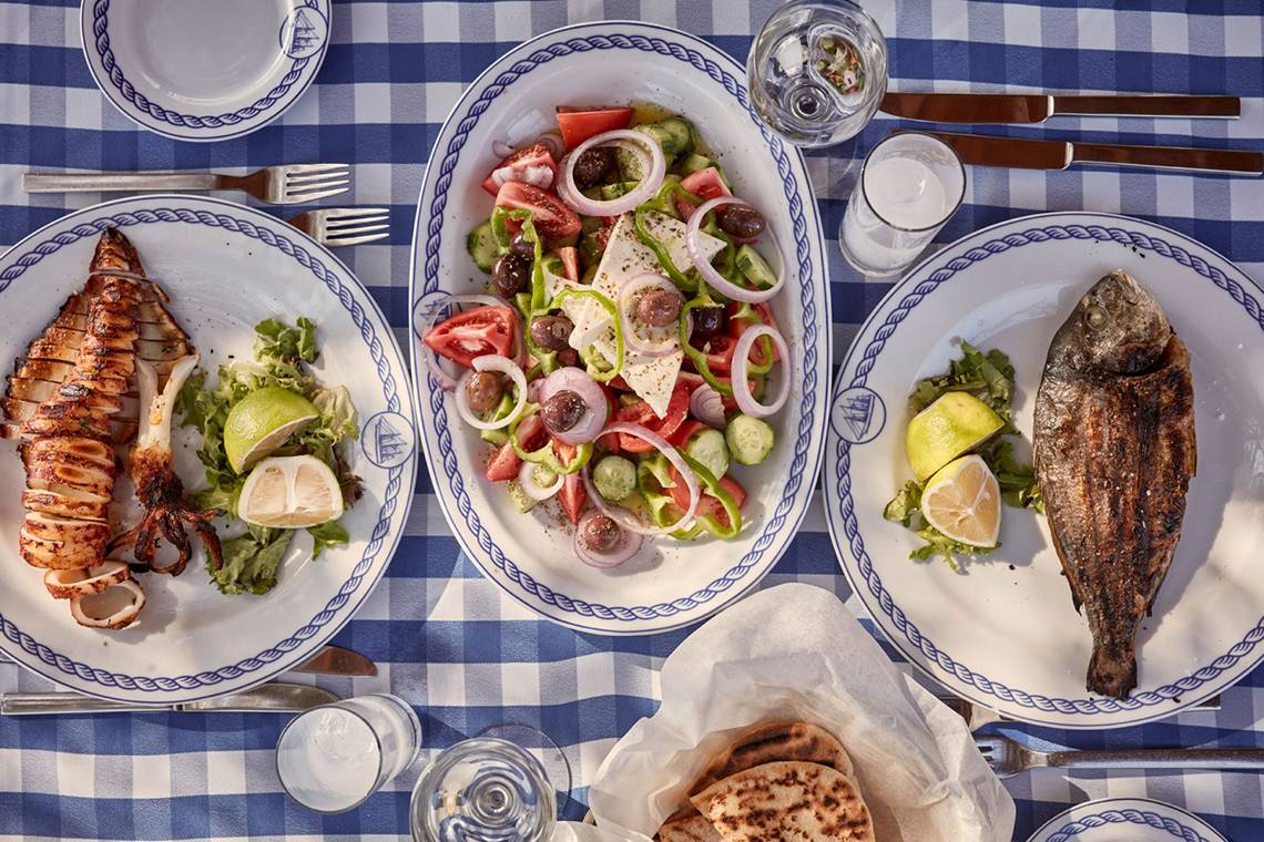 Griechisches Essen auf blauem Karotisch, inklusive gegrillter Tintenfisch, Salat und gebackener Fisch.