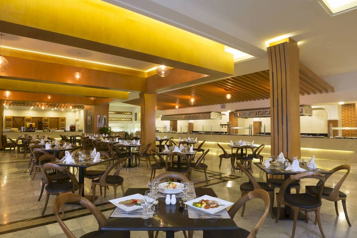 Innenansicht des Restaurants im Pickalbatros Aqua Park Resort - Sharm, mit gedeckten Tischen und Stühlen, gelben Deckenleuchten und einer Theke im Hintergrund.