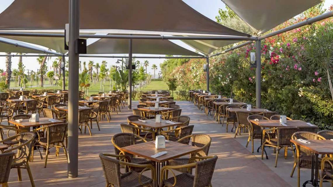 Terrasse des Royal Seginus Hotels mit vielen runden Tischen und Stühlen unter Sonnensegeln.