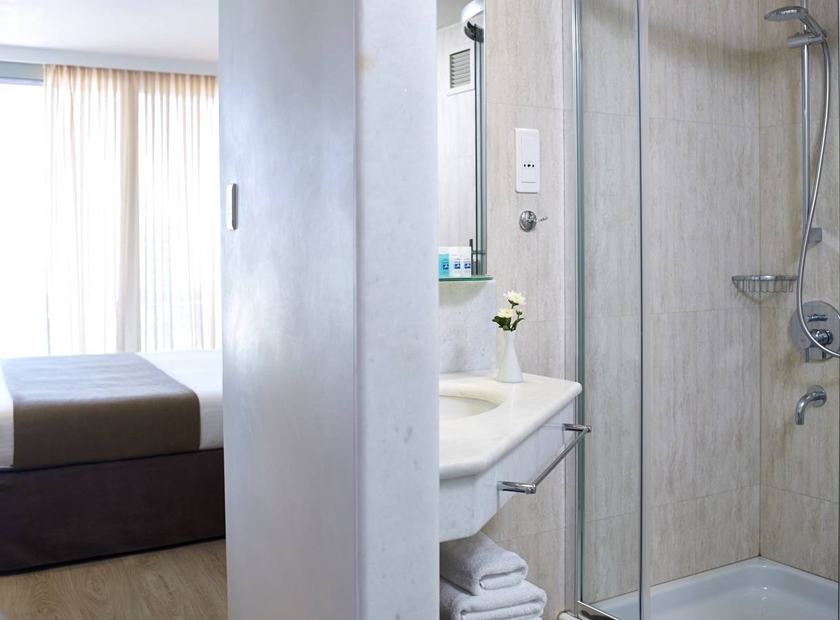Badezimmer im Kriti Beach Hotel mit Dusche, Waschbecken und Bett im Hintergrund.