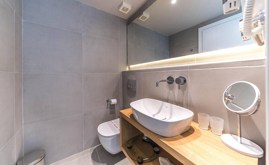 Badezimmer im Adele Beach Hotel mit grauen Fliesen, Toilette, Waschbecken und Spiegeln.