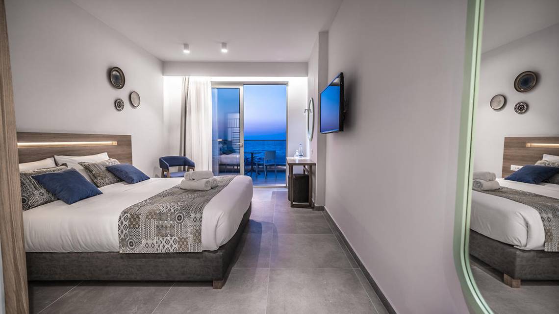 Innenansicht eines Hotelzimmers im Adele Beach Hotel mit einem Doppelbett, einem Fernseher und einem Balkon.