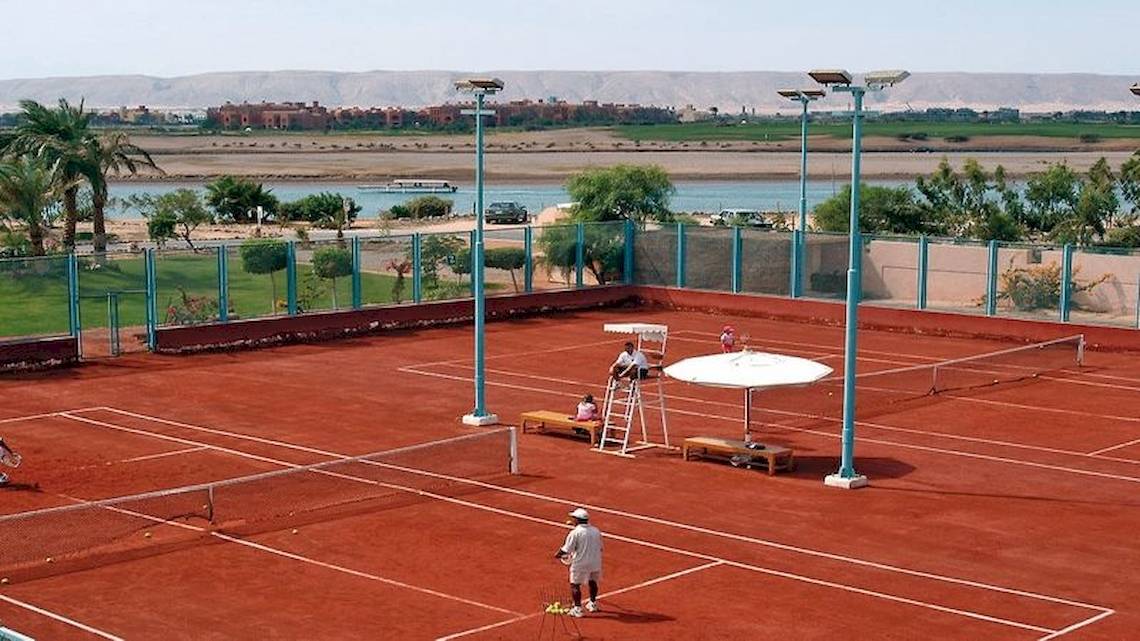 Tennisplatz im Movenpick Resort & Spa El Gouna mit Spielern, einer Tribüne und einem See im Hintergrund.