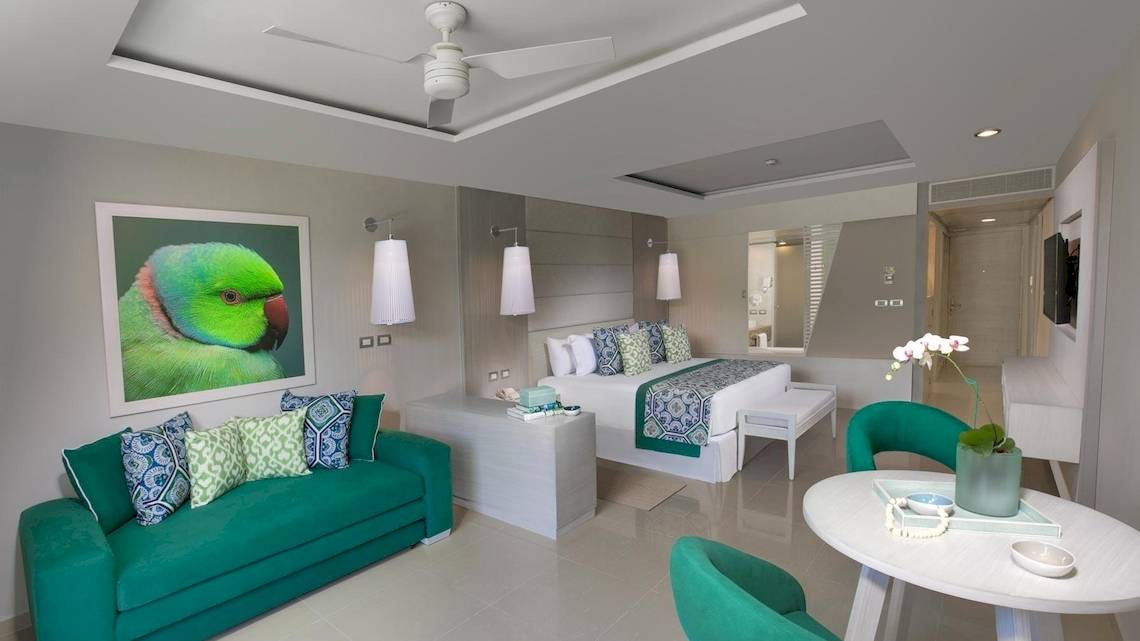 Innenansicht eines Zimmers im Grand Sirenis Riviera Maya Resort & Spa mit grünem Sofa, Bett, Tisch und Kunstwerk an der Wand.