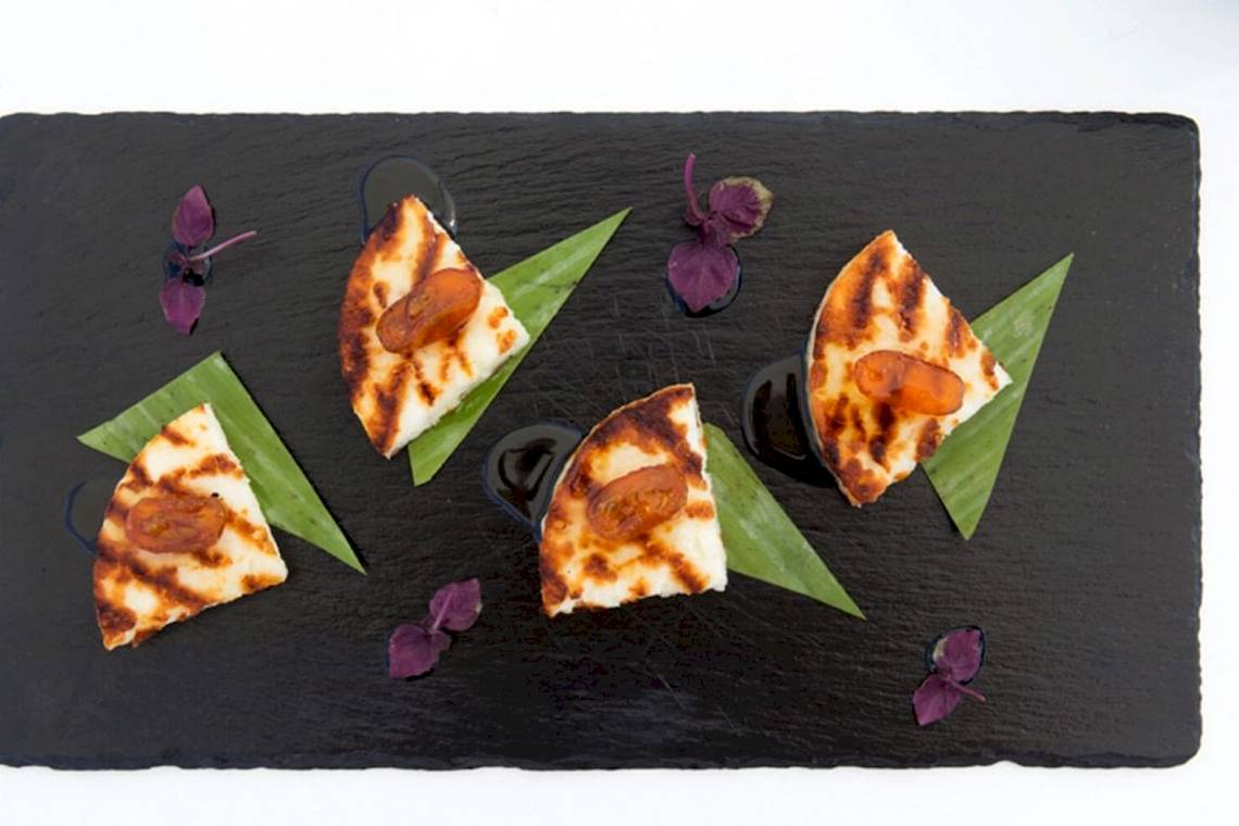 Gegrillter Käse mit Aprikosen auf einer schwarzen Platte mit grünen Blättern und violetten Blüten.