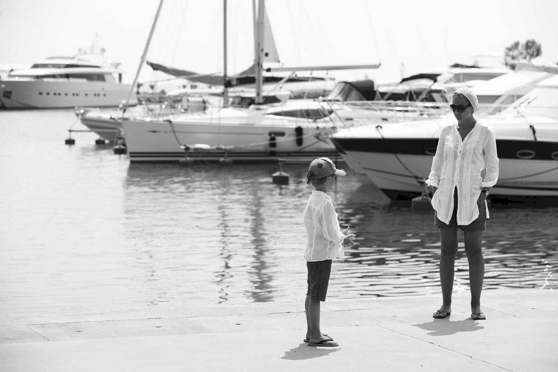 Schwarz-Weiß-Foto von einem Hafen mit mehreren weißen Booten und zwei Personen, einer Frau und einem Kind, die am Ufer stehen.