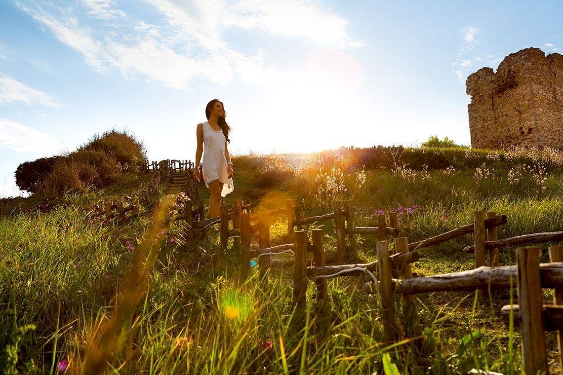 Eine Frau in einem weißen Kleid steht in einem Feld mit einem alten Steinturm im Hintergrund.