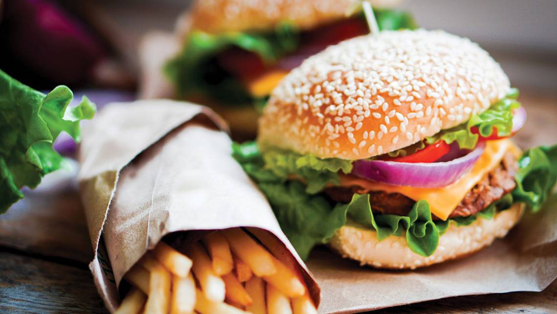Zwei Burger und Pommes auf braunem Papier, eines mit Zwiebeln und Salat, das andere mit Tomaten und Salat.