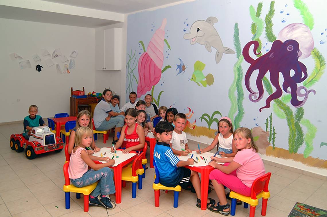 Kinder sitzen an Tischen in einem Raum mit Meerestier-Wandmalerei, darunter ein Delphin und eine Muschel.