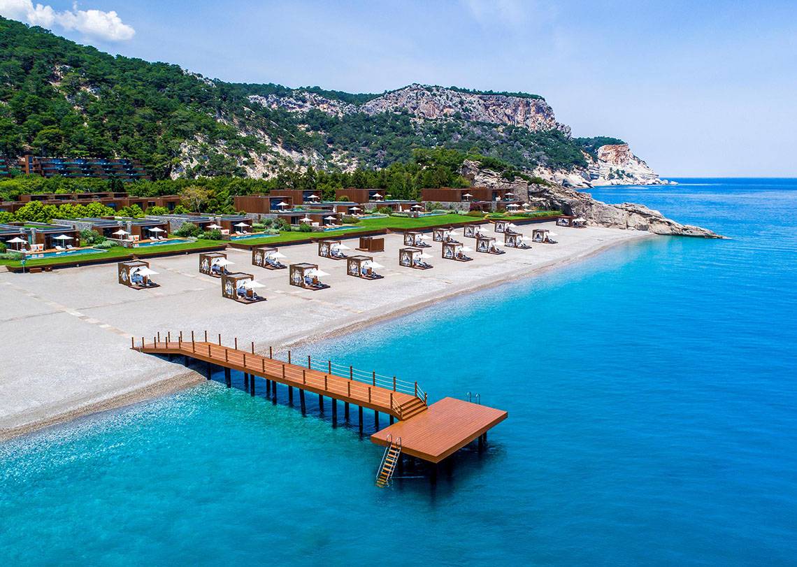 Luftaufnahme des Maxx Royal Kemer Resorts mit weißen Sonnenliegen auf dem Strand und blauem Meer.