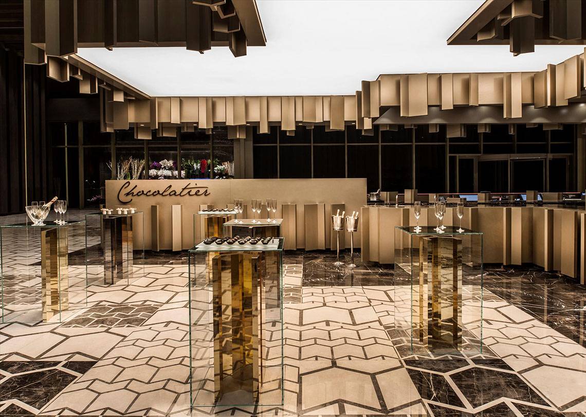 Ein eleganter Bereich im Maxx Royal Kemer Resort mit Glastischen, goldenen Säulen und einer Theke mit dem Wort Chocolatier.