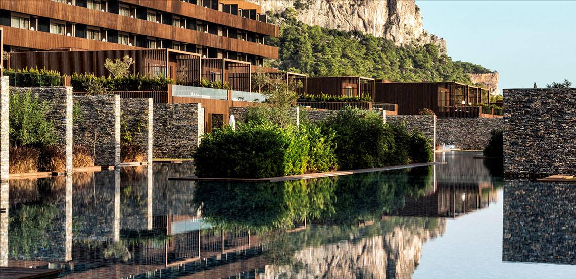 Außenansicht des Maxx Royal Kemer Resorts mit steinerner Einfriedung und Pool, dahinter ein felsiger Hügel.