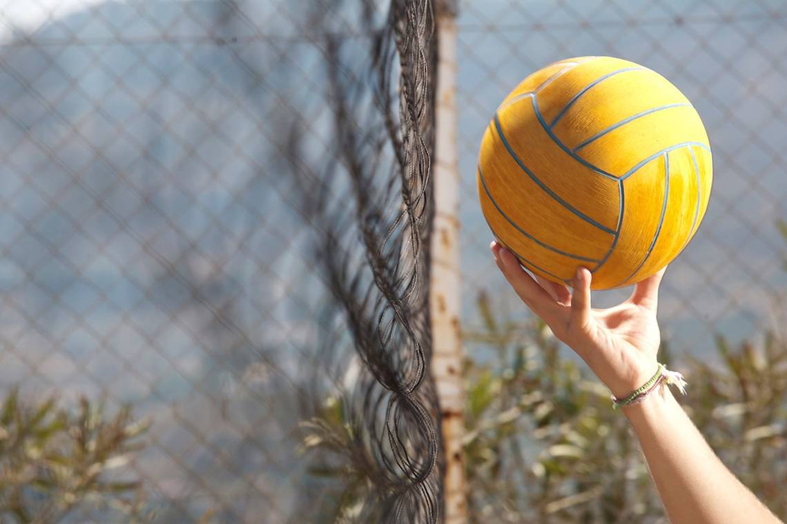 Hand hält einen blauen Wasserball vor einem Netz im Freien.